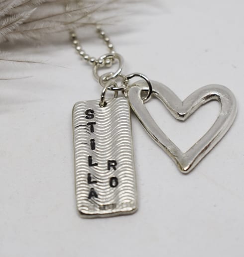 halsband med hjärta och platta i silver med texten stilla ro på vit botten med fjäder i bakgrunden