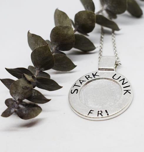 silverhalsband med texten STRAK, UNIK, FRI med vit bakgrund och grön kvist bredvid